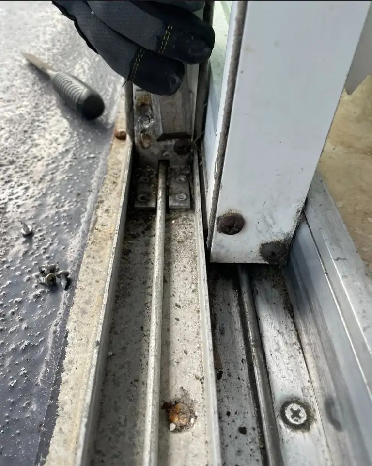How to clean sliding glass door tracks: a dirty aluminum sliding door bottom track with built-up grime and a rust-stained screw, a gloved hand at the door panel