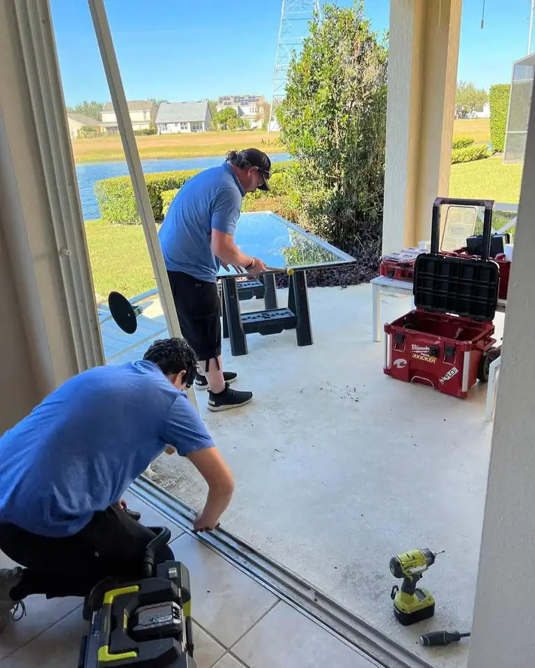 Sliding glass door off track for repair: a Stuck Door technician works on the empty bottom track while a second technician handles the removed glass panel on sawhorses